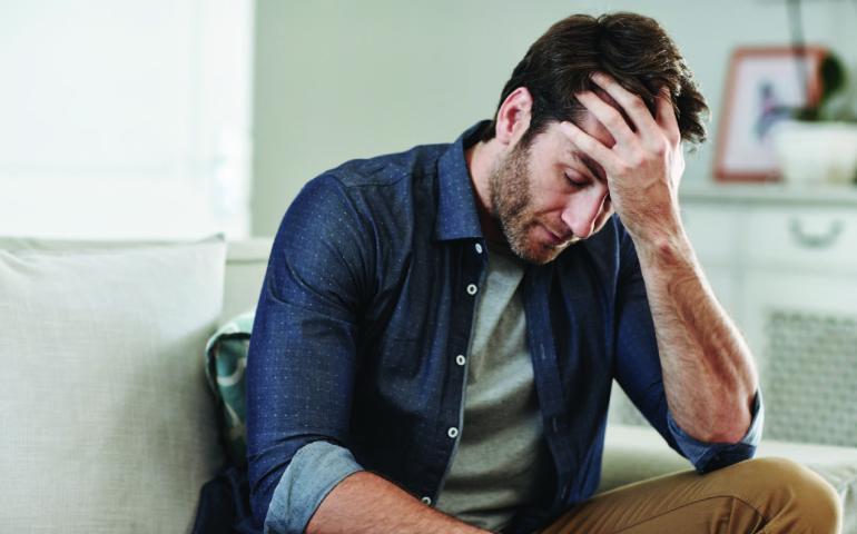 Man seated on couch with his hand on his forehead looking fatigued and stressed